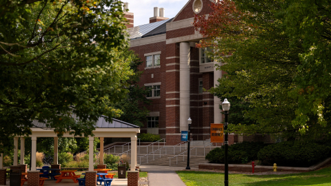a scenic shot of campus features a covered patio with orange blue chairs, with Rooke Science Center in the backdrop and lush green foliage.