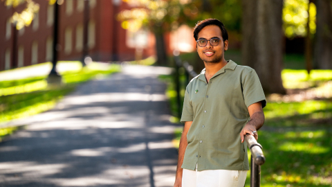 A Bucknell student wearing a green shirt stands outside on a sunny day with trees and a walking path in the background.