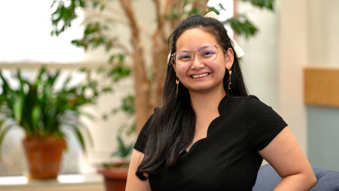 Angel Eunisse Yanga wears a black short-sleeved shirt and smiles in a room where there are potted plants in the background.