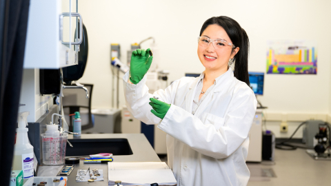 My Ly wears a white lab coat, safety goggles and green gloves while holding lab equipment in a lab setting.