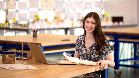 Katie Raso '28 sits in a classroom at a desk with a laptop and papers in front of her and smiles.
