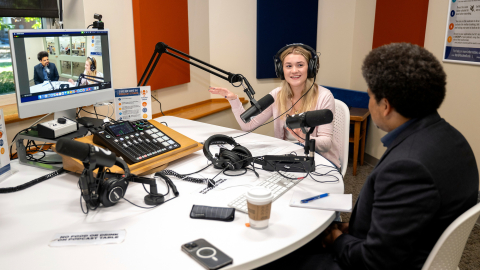 Rayna Tyler '27 wears headphones and sits at a table with a microphone and computer screen while speaking to a guest, who has his back to the camera.