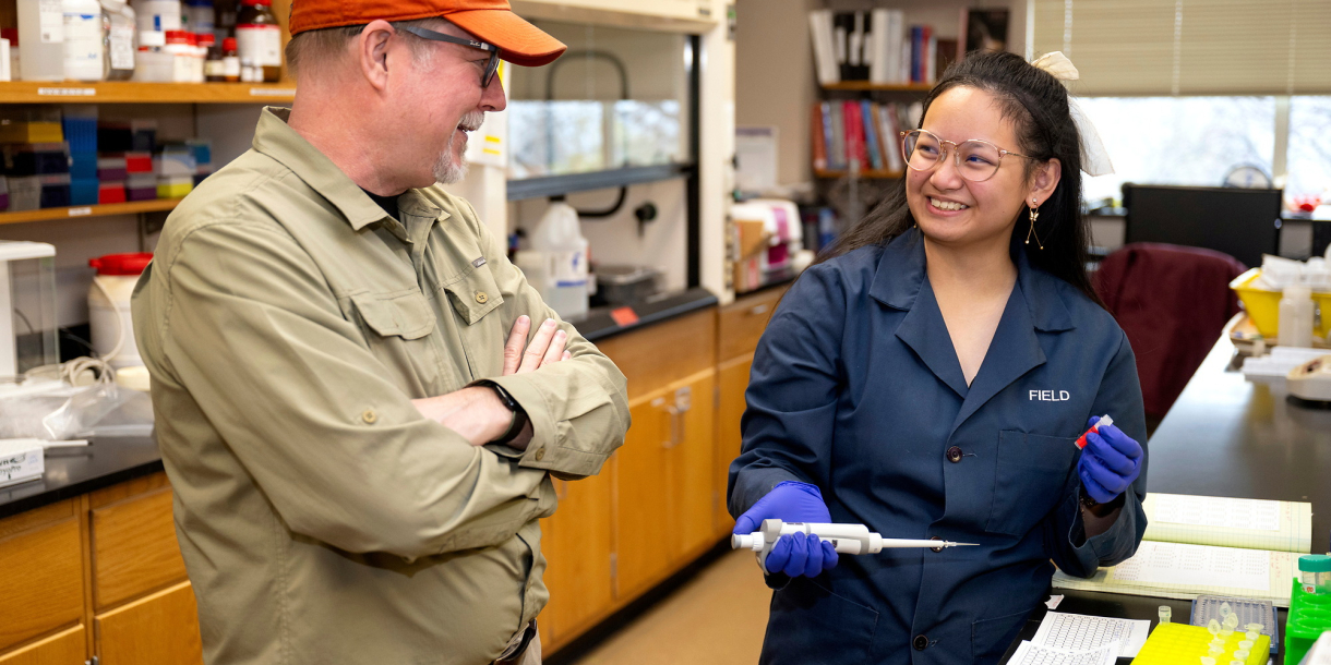 Angel Eunisse Yanga is smiling in a campus lab space wearing a blue lab coat while talking with Professor Ken Field.