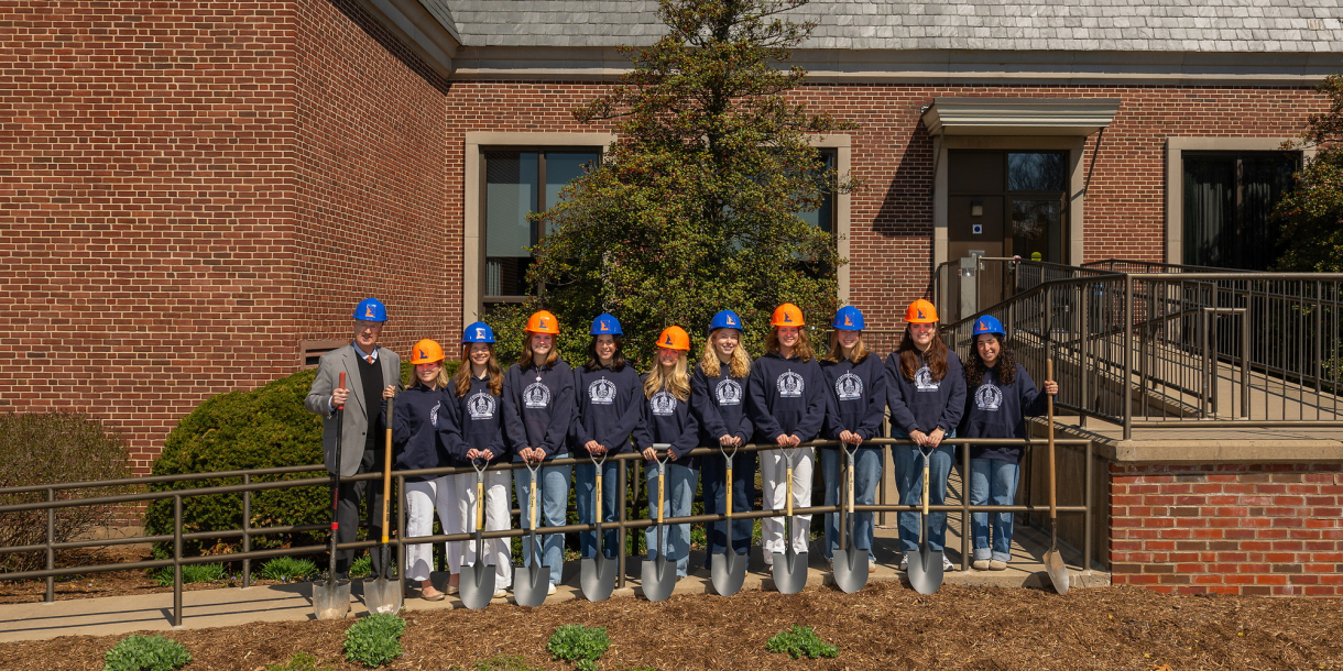 a group of students wearing orange and blue hard hats and blue hooded sweatshirts stand in a line together with President John Bravman outside the ELC.