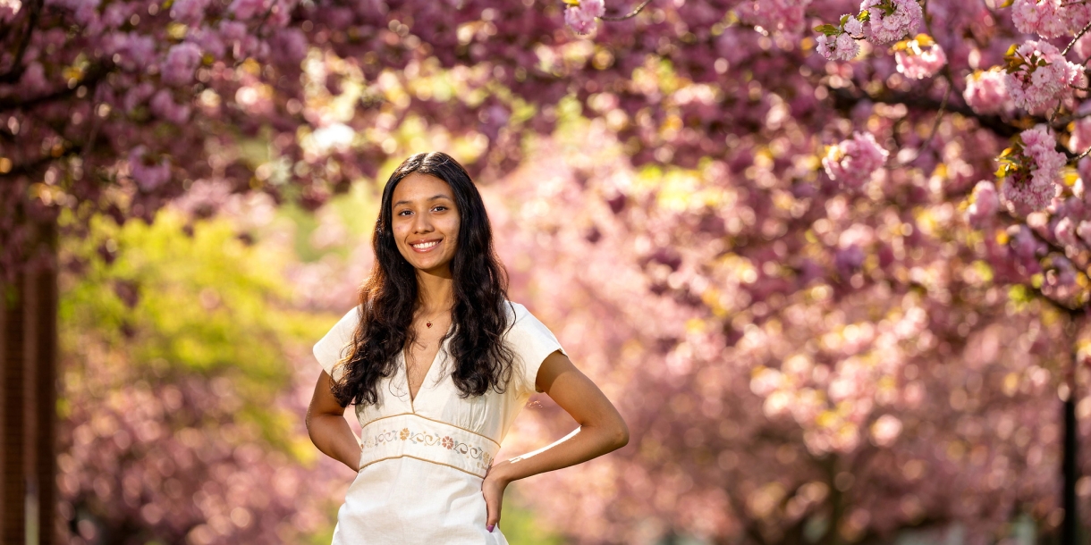 Gabby Diaz '25, in a white dress, poses with her hands on her hips in front of cherry blossom trees.