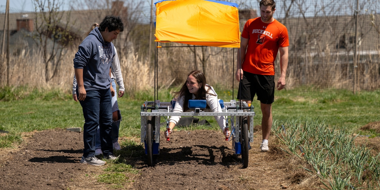 A student lies down, facing the ground, in a cart-like device that allows for planting and weeding of crops at the Farm. Three classmates walk along side the cart-like machine.
