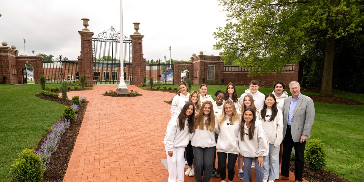 A group of students stands with President John Bravman at the bottom of a wide, brick pathway that leads through the Christy Mathewson Gates on campus.