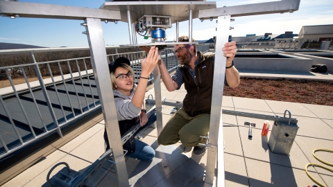 A female student is looking under a metal frame at a mechanism that drives a vertical wind turbine. A male professor kneels across from her.