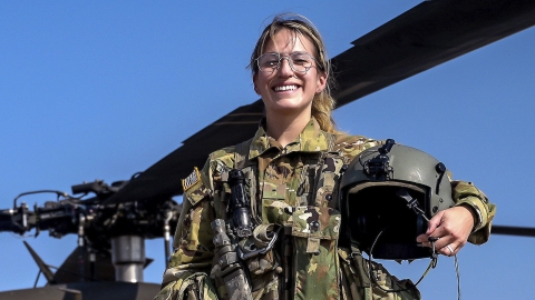 Nicole Vacas-English is in a camo flight suit holding a helmet and smiling with a helicopter behind her