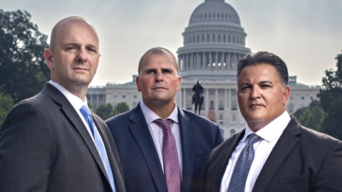 Three men in suits and serious expressions look at the camera with the US Capitol Building in the background