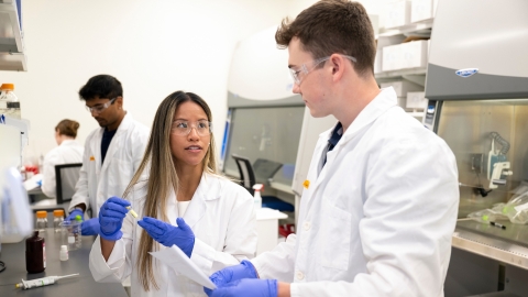 Professor Olivia Boerman and Connor Kozick &#039;26 are wearing white lab coats and purple gloves in a lab setting.