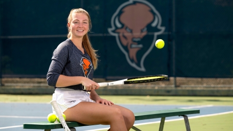 Anna Lajos '25 bounces a tennis ball on a racquet, sitting on a bench beside a Bucknell tennis court.