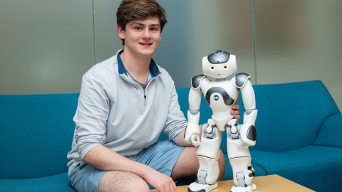 Sean O&#039;Connor sits on a blue couch and smiles with a robot standing on a table next to him..
