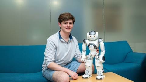 Sean O'Connor sits on a blue couch smiling next to a robot. 