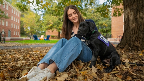 Emma Feld sits on a blanket of leaves on the ground with a young black lab puppy.