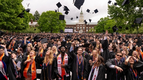 Celebratory members of the Class of 2024 toss their graduation caps in the air and cheer and smile.