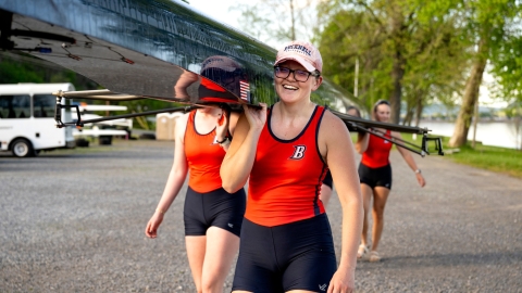 Kona Glenn is wearing an orange and blue rowing uniform and a pink Bucknell baseball cap as she smiles and carries a boat along a riverbank.