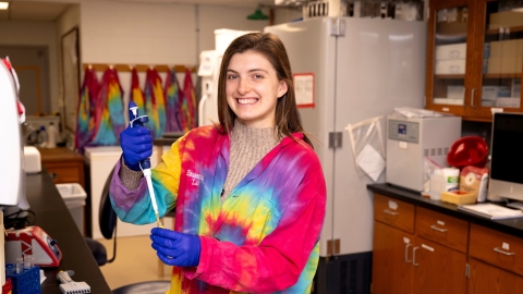 Meghan Catherwood &#039;25 wears a bright, multi-colored tie-dyed lab coat and blue gloves while smiling and holding a pipette in a lab on campus.