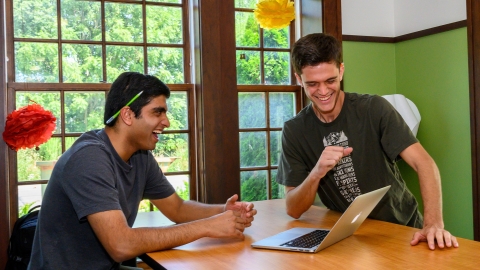 Two male students smile gathered around a laptop on a table.