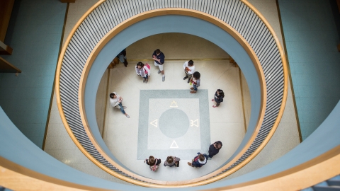 Overhead view of a spiral staircase with people standing in a circle on the ground floor. The stairway's curves frame the scene, creating a concentric pattern.