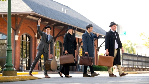 Four people dressed as salesmen cross an intersection in front of a trainstation.