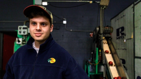 Ben Fink stands with a conveyer belt with apples on it behind him.