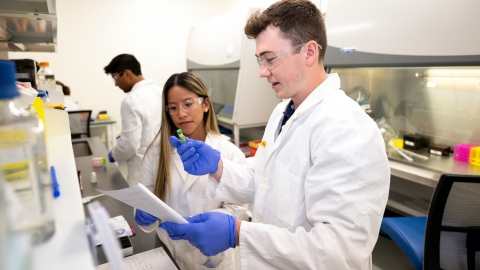 Professor Olivia Boerman and Connor Kozick &#039;26 are wearing white lab coats and purple gloves in a lab setting.