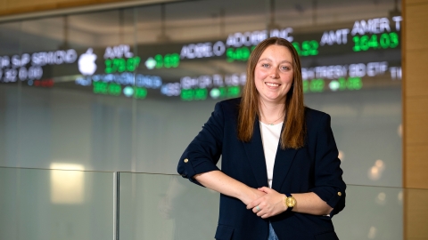Caitlin Yant &#039;24 stands and smiles in the Moriatry Investment Center in Holmes Hall with a stock ticker behind her.