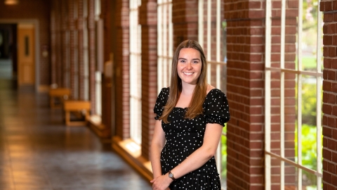 Kendall Robertson wears a black dress and smiles while standing in a hallway with brick columns and windows behind her.