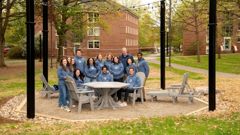 President John Bravman and Bucknell Student Government members from the Class of 2023 gather at a table on an octantal-shaped pit, with four pillars and stringed lights overhead.