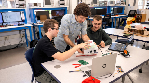 Two students sit at a table in a makerspace with a staff member in between them as they look at pieces of technical hardware together.