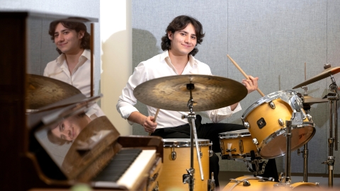 Darren Mayrowetz '27, dresed in a white collared shirt and black pants, plays a drum set next to a piano.