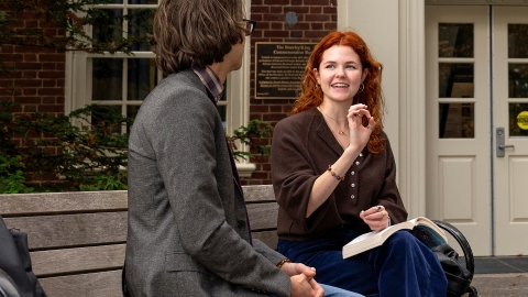 Hailey Drapcho sits on a bench outside an academic building and is in a dynamic conversation with a faculty member who you see the back of.