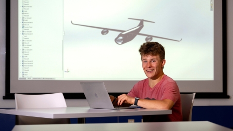 Kade Davidheiser sits at a table on his laptop with an image of an airplane model projected on the screen behind him