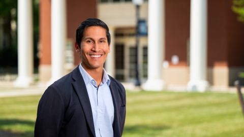 Professor Karlo Malaga stands on campus and smiles, a building with columns is in the background