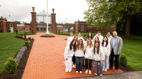 A group of students stands with President John Bravman at the bottom of a wide, brick pathway that leads through the Christy Mathewson Gates on campus.