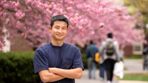 Tobey Kim wears a blue T-shirt and smiles on campus with cherry blossom trees behind him.