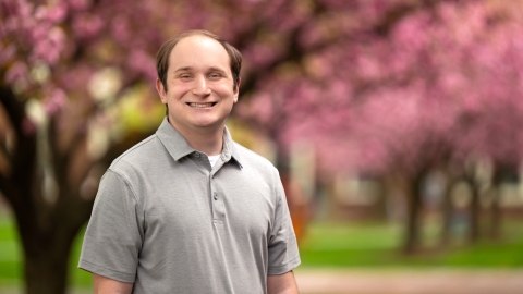 Zane Hensal wears a gray short-sleeved colored shirt and smiles on campus with cherry-blossom trees behind him.
