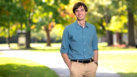 Sam Gutekunst is wearing a blue shirt and tan pants and stands and smiles on campus with sunlight shining through green leaves behind him. 