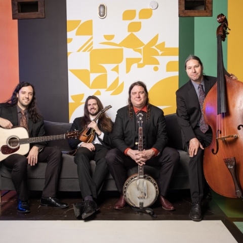 Members of Henhouse Prowlers sit on a couch facing the camera, each holding their instrument