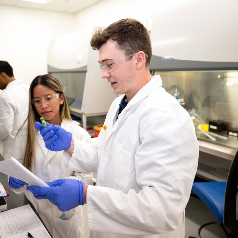 Professor Olivia Boerman and Connor Kozick &#039;26 are wearing white lab coats and purple gloves in a lab setting.