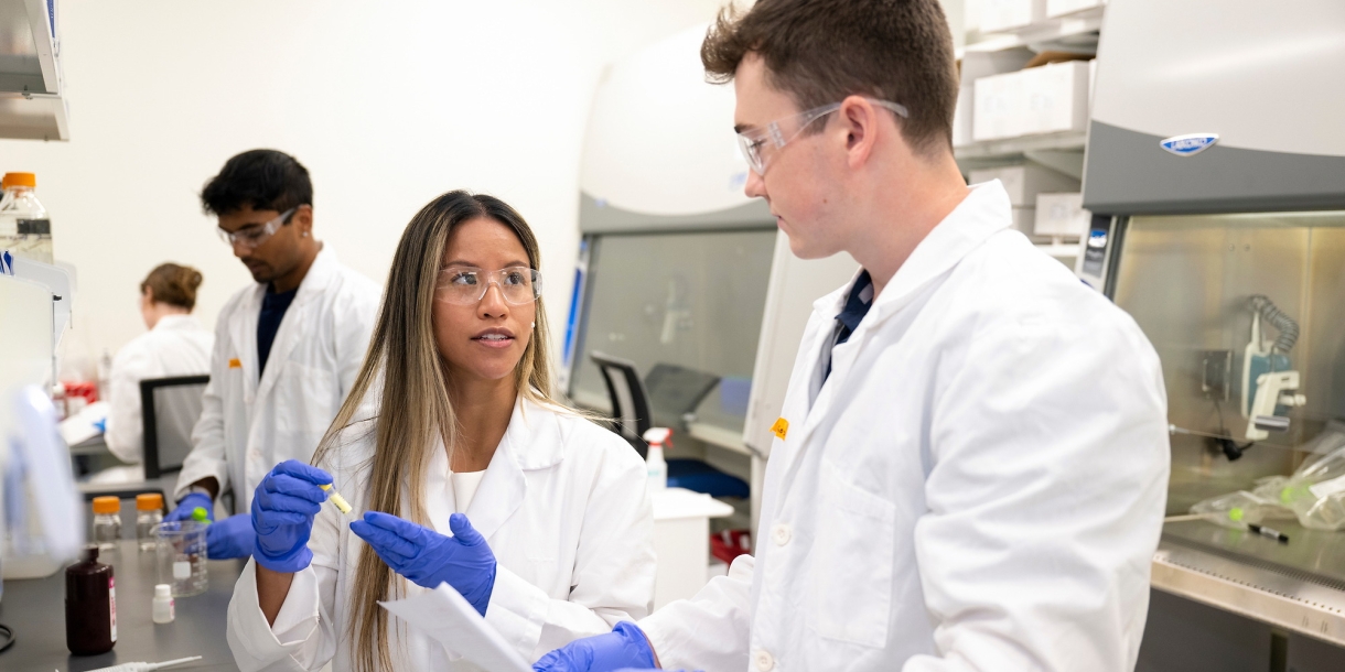 Professor Olivia Boerman and Connor Kozick &#039;26 are wearing white lab coats and purple gloves in a lab setting.