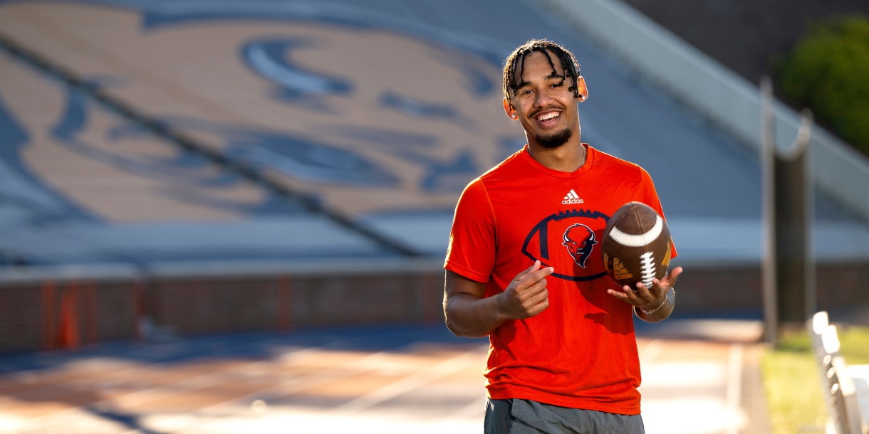 Michael Hardyway '25 wears an orange Bucknell shirt and holds a football as he smiles in Christy Mathewson Memorial Stadium.