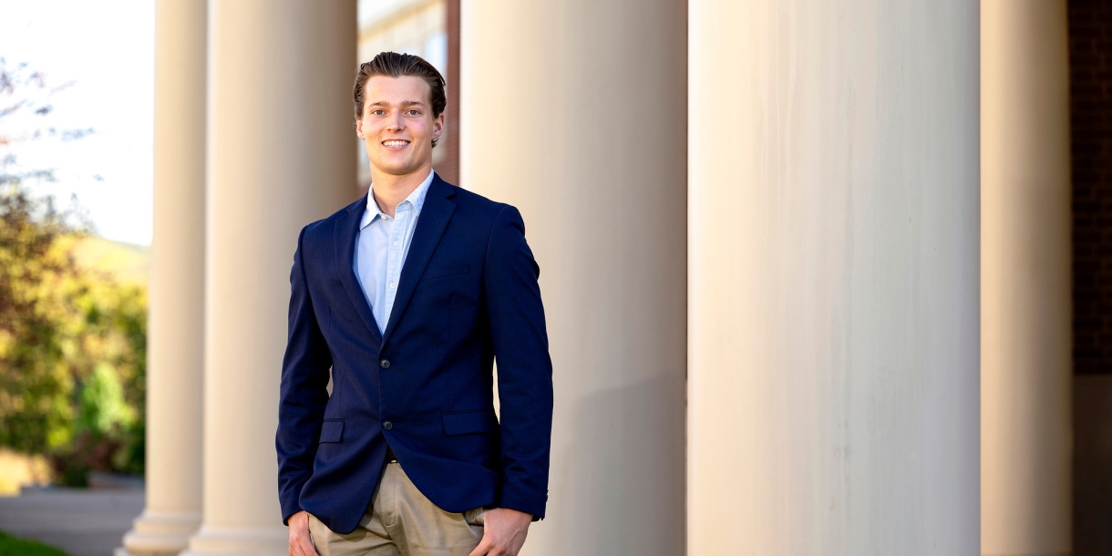 Nick Sperduto &#039;24 wears a navy blazer and tan pants and smiles while standing in front of columns on campus.