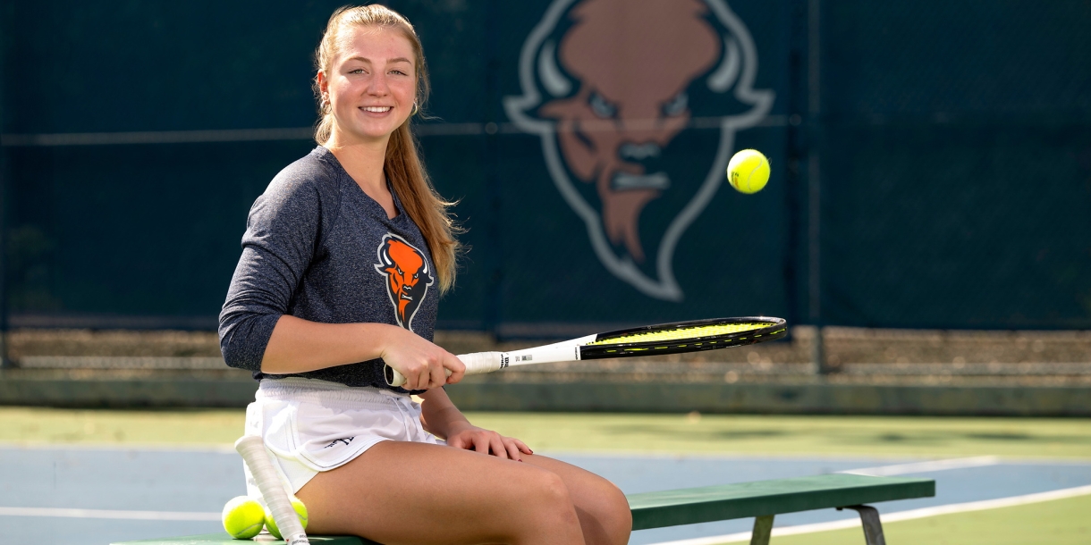 Anna Lajos '25 bounces a tennis ball on a racquet, sitting on a bench beside a Bucknell tennis court.