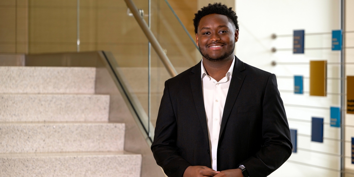 Jerimaha McClain wears a dark suit and white dress shirt and stands at the base of a stairway in Holmes Hall.