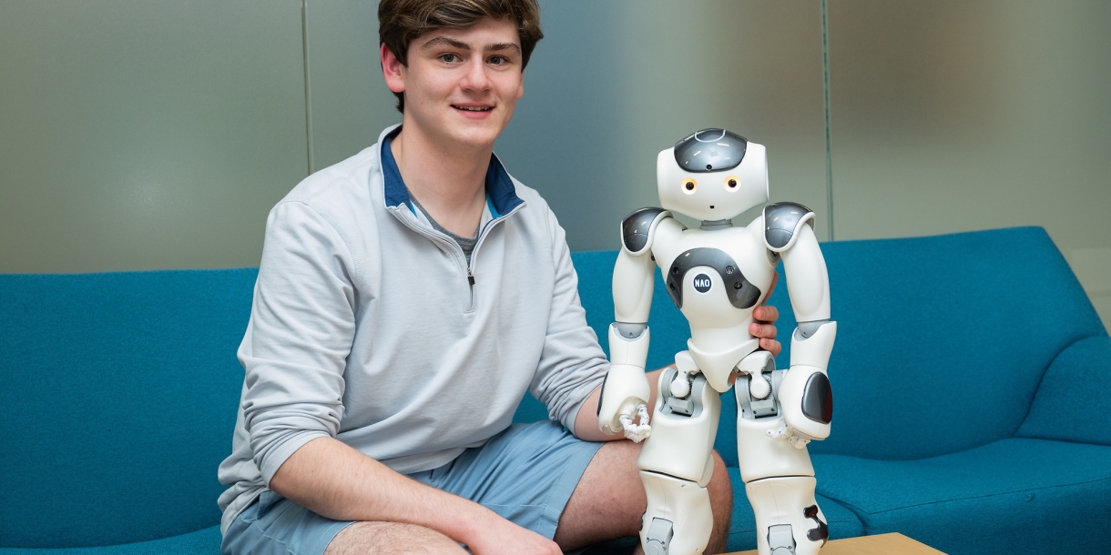 Sean O&#039;Connor sits on a blue couch and smiles with a robot standing on a table next to him..