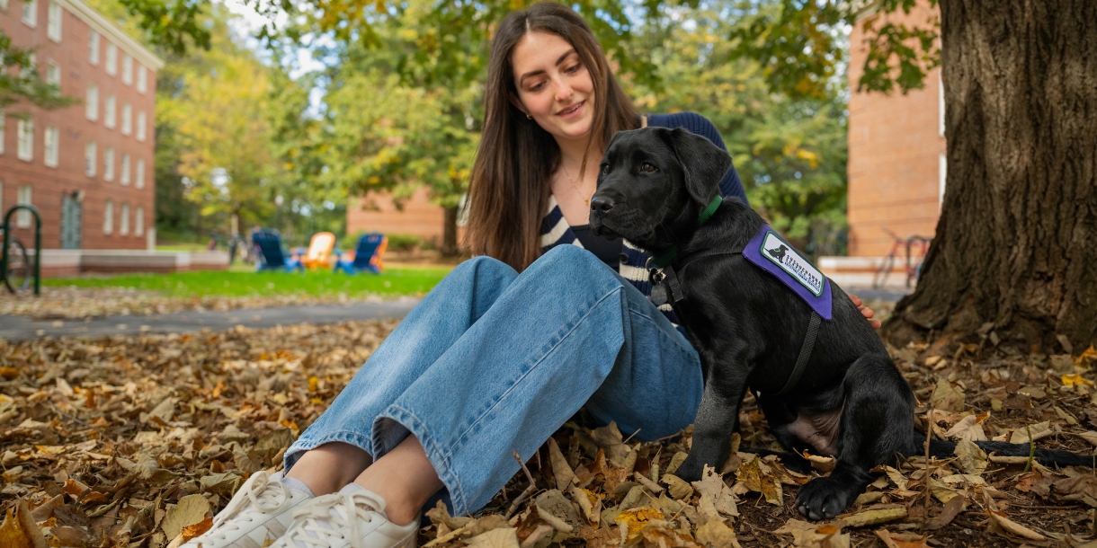 Emma Feld sits on a blanket of leaves on the ground with a young black lab puppy.