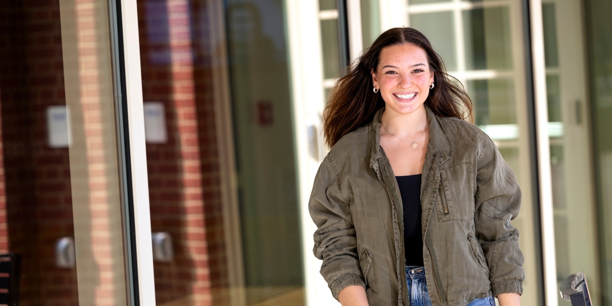 Elizabeth Malley wears a tan jacket and jeans and walks in front of a building on campus with a very big smile.