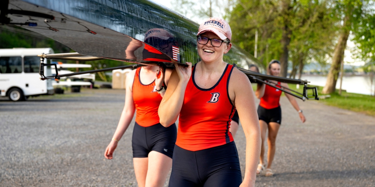 Kona Glenn is wearing an orange and blue rowing uniform and a pink Bucknell baseball cap as she smiles and carries a boat along a riverbank.